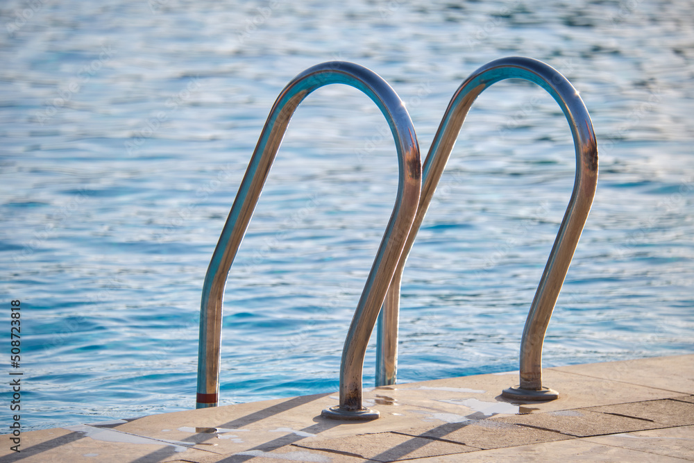 Close up of swimming pool stainless steel handrail descending into tortoise clear pool water. Accessibility of recreational activities concept Close up of swimming pool stainless steel handrail descending into tortoise clear pool water. Accessibility of recreational activities concept