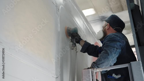 Close-up of a paint line worker in a respirator sanding the car wall before finishing painting using a mechanical sander. Dusty work of a painter in the paint shop of a wagon plant