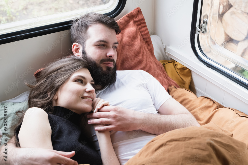 Young just married couple is traveling in travel van. Romantic atmosphere of relaxation. Road trip around country for weekend. Caucasian man and woman lying in bedroom in camper. Millennial generation