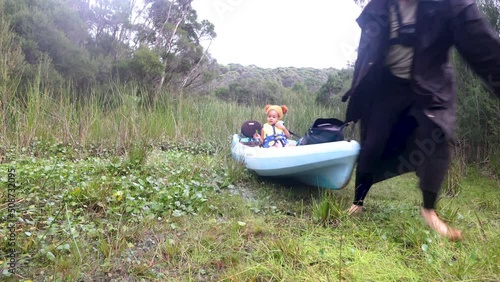 A man in a rain jacket drags a kayak through swamp land with his daughtet sitting in the kayak. photo