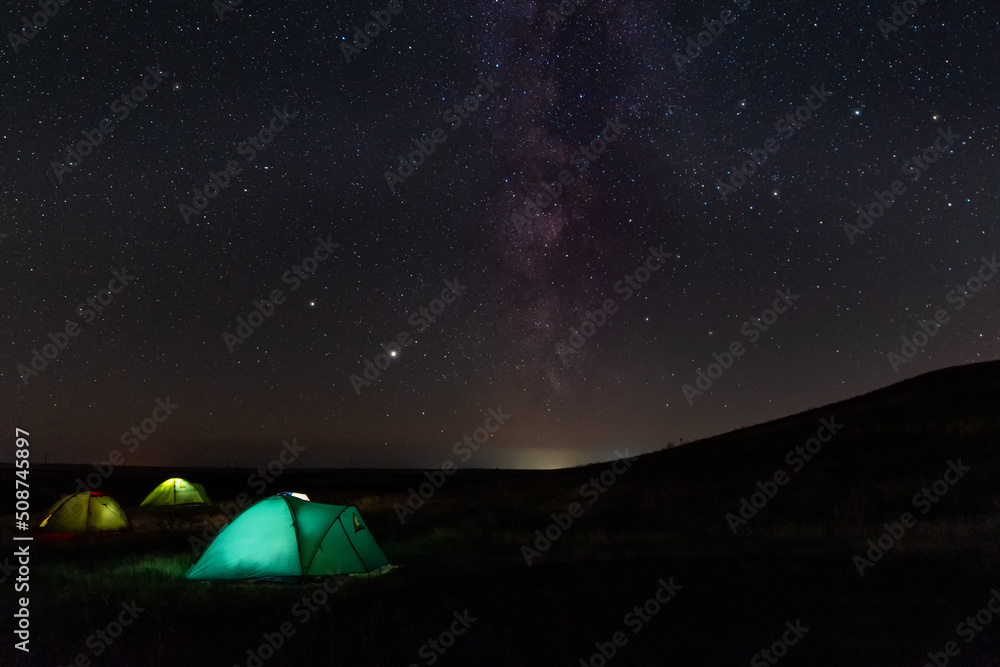 Travel and camping concept - three glowing camping tents at night under a starry sky with milky way and setting sun. Illuminated blue camping tent under stars at night