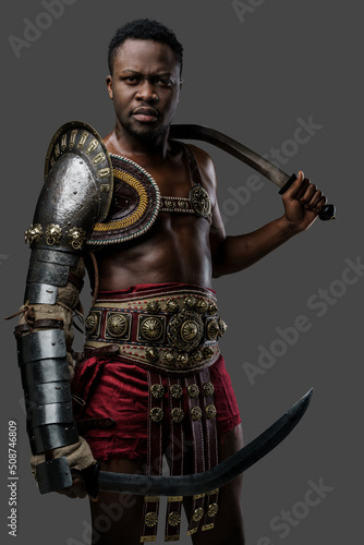 Portrait of serious african arena fighter dressed in armor holding twin swords against grey background.