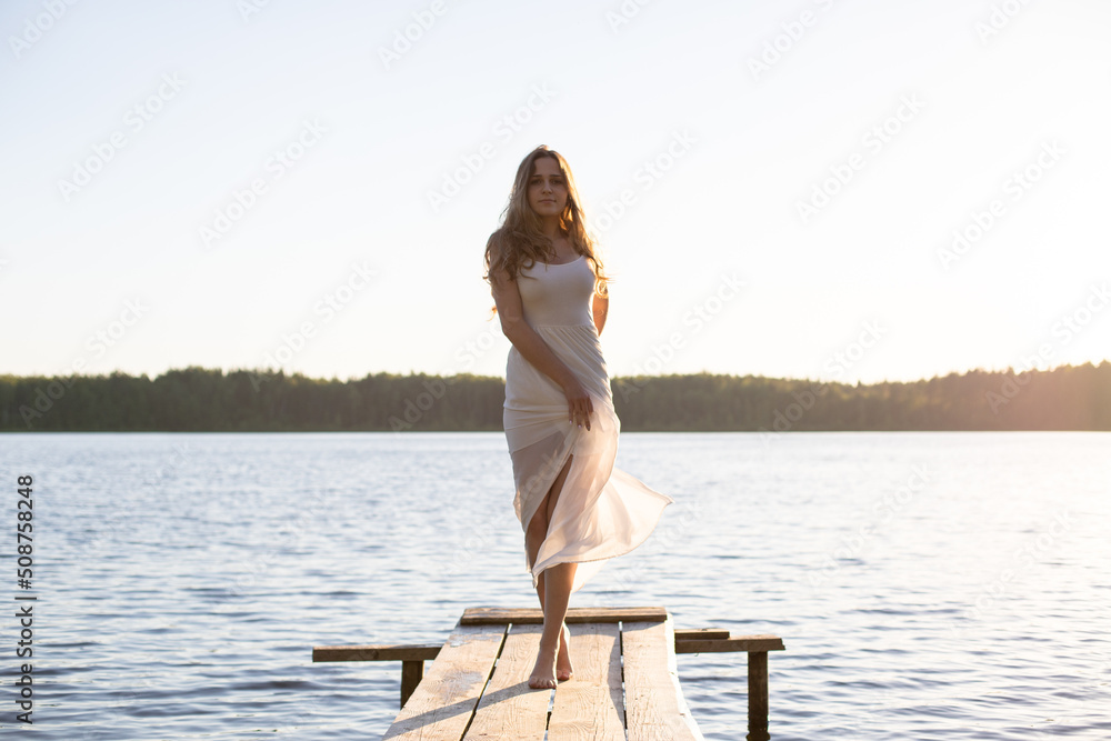 Beautiful Russian girl dressed in a white dress, walking along a wooden pier on the bank of a river or a lake Beautiful Russian girl dressed in a white dress, walking along a wooden pier on the bank of a river or a lake