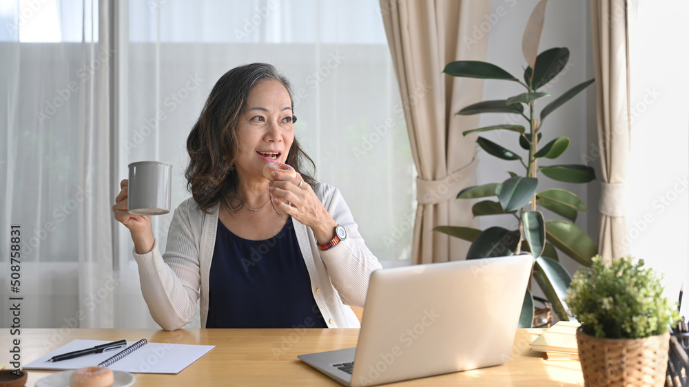 Satisfied middle aged woman holding coffee cup and using computer laptop in bright modern living room Satisfied middle aged woman holding coffee cup and using computer laptop in bright modern living room