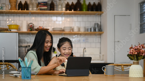 Asian mom helping her little daughter doing homework while siting together at home kitchen