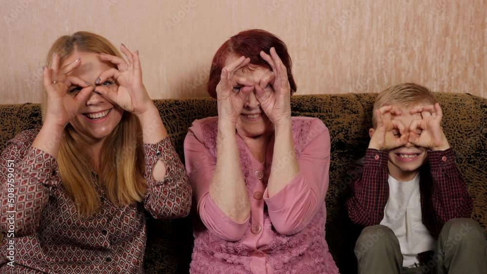 A great-grandmother with her daughter and grandson are having fun at home sitting on the couch, they make glasses with their hands and look at the camera. A great-grandmother with her daughter and grandson are having fun at home sitting on the couch, they make glasses with their hands and look at the camera.