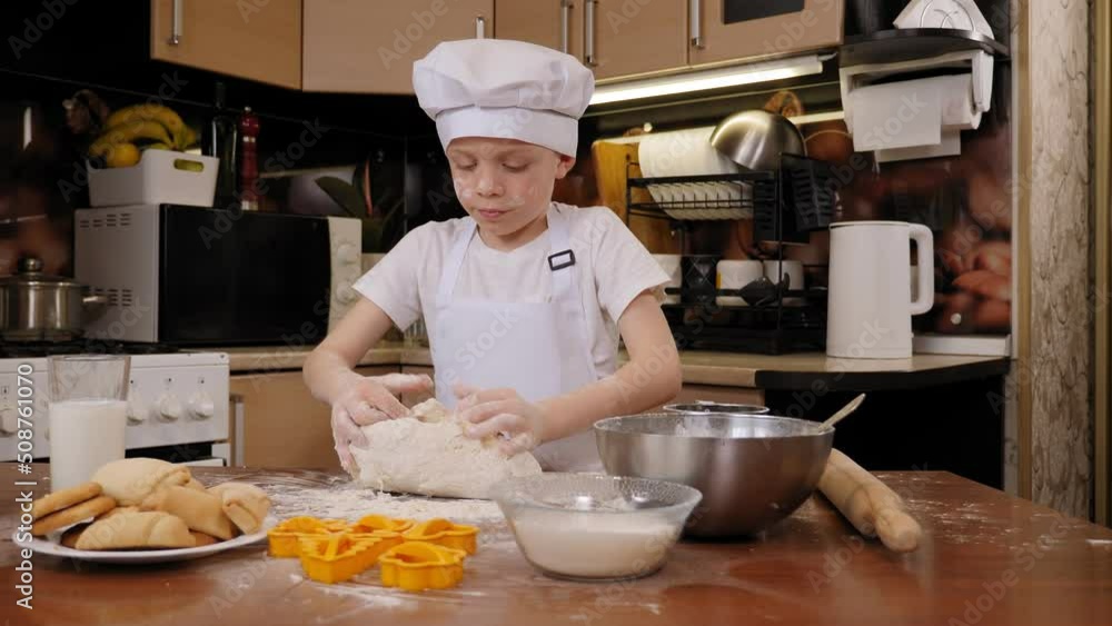A little cheerful boy with a face covered in flour kneads the dough at home in the kitchen. A cute child is preparing cookies. A little cheerful boy with a face covered in flour kneads the dough at home in the kitchen. A cute child is preparing cookies.