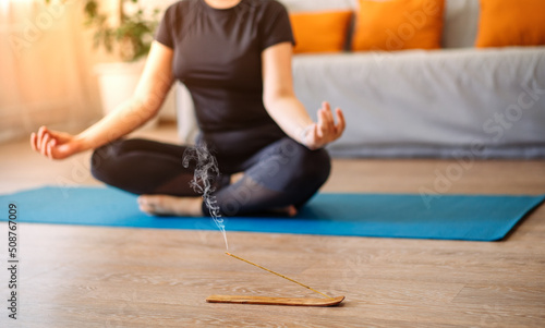 A woman meditates in the lotus position on a turquoise yoga mat on the wooden floor of the house in the living room. Focus on the incense stick and the smoke.