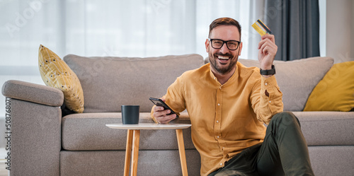 Portrait of cheerful man with credit card using mobile phone in living room at home photo