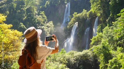 Young woman traveler make photos for social media of amazing exotic waterfalls and tropical nature on her smartphone camera. Happy female tourist with backpack in travel. Wanderlust, tourism concept. photo