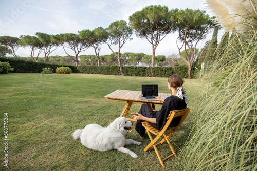 Young caucasian woman watching something on laptop computer and drinking tea while sitting at wooden table on green mediterranean lawn. Concept of weekend  rest and remote work