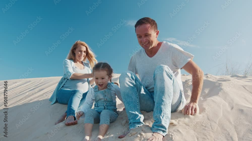Family spend time together. Young family of father, pregnant mother and toddler girl enjoy playing together on the sandy open space