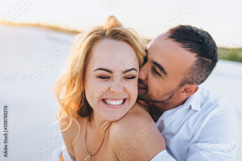A tanned young caucasian bearded guy kisses a beautiful fun smiling blonde woman against the backdrop of the setting sun. Desert, sandy beach, rest and relaxation. Honeymoon concept.