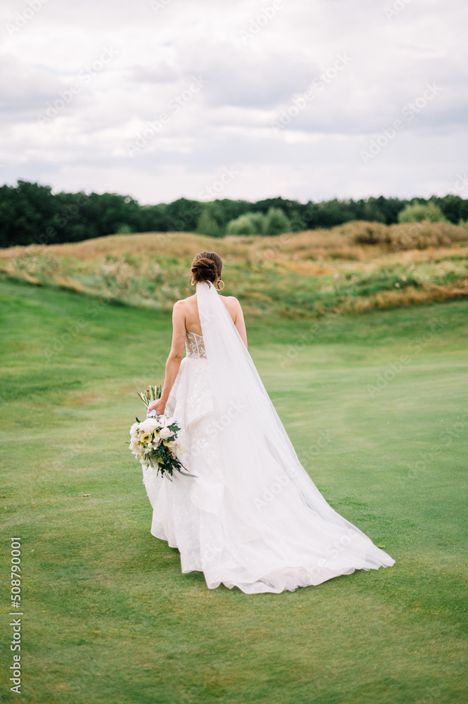 Portrait of beautiful bride in white wedding dress with modern hairstyle walking on green golf course , back view. Wedding concept