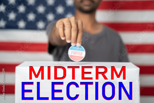 Concept of 2022 American Midterm Elections showing by placing I voted sticker on ballot box with Midterm Election sign in front of US flag photo