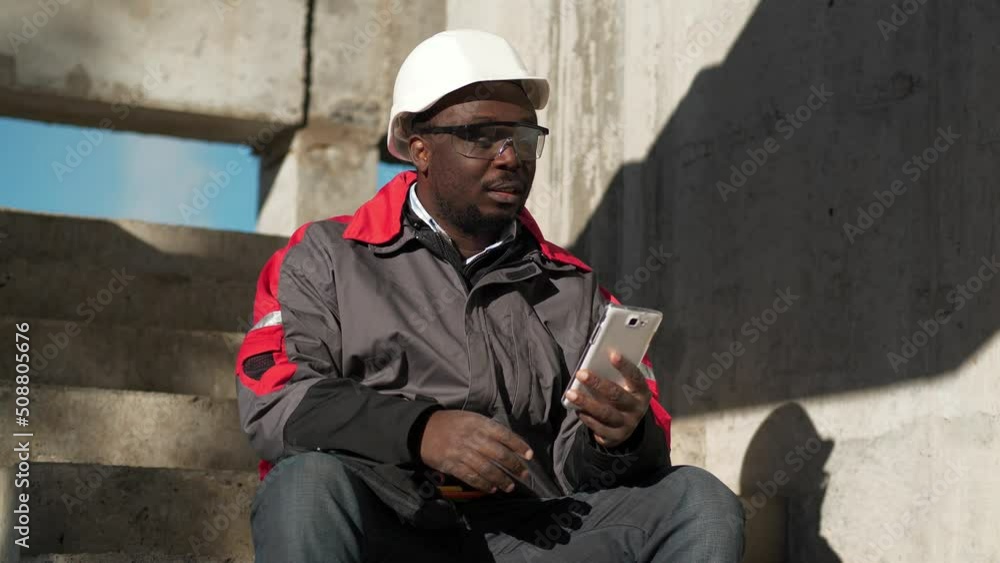 Slow Motion African american builder sits on the stairs at construction site and talks on smartphone by video link Slow Motion African american builder sits on the stairs at construction site and talks on smartphone by video link