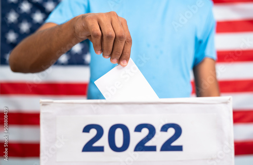 Man placing ballot paper into 2022 ballot box in front of american flag - concept of 2022 midterm US election, voting and democracy