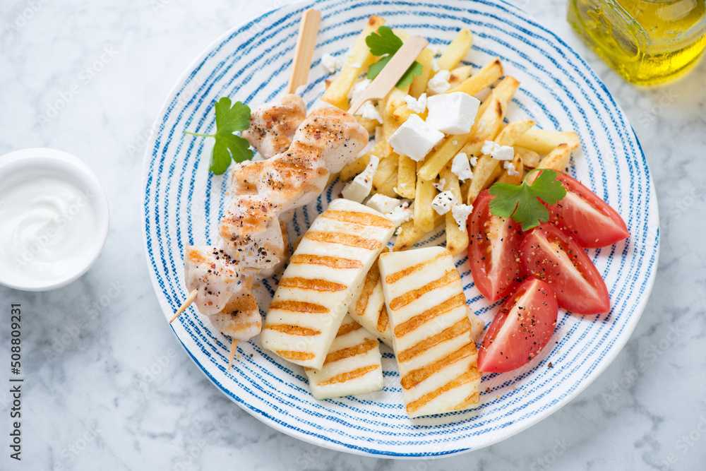 Blue and white plate with grilled halloumi, souvlaki, greek-style fries with feta and tomatoes, studio shot on a light-grey marble background