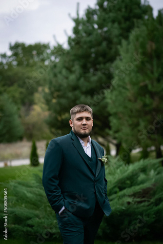 Stylish man in a green suit in front of a pool and a house in a green park. A garland of bulbs in a blur.