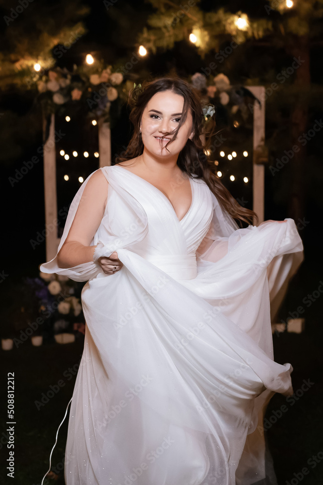 Lush girl in a white wedding dress. Stylish charming and cheerful bride. Green park with light bulbs in the background.