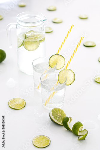  On a white table is a glass jug of lime lemonade, next to it are two glasses filled with lemonade and lots of limes.