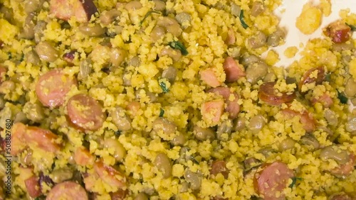 Cooking and mixing with a spoon the Northeastern Feijao Tropeiro, brazilian dish made with cuscuz, string bean, sausage, bacon, chive and curd cheese. Zoom out effect.