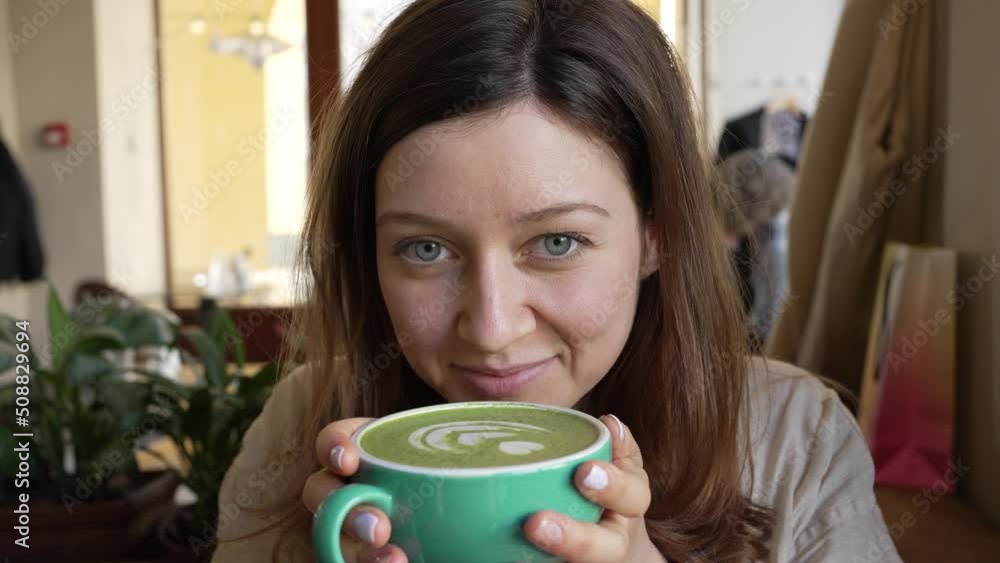 Young woman in cafe takes sip of matcha latte from green cup. Woman tastes trendy matcha tea and pleasantly smiles looking at camera Young woman in cafe takes sip of matcha latte from green cup. Woman tastes trendy matcha tea and pleasantly smiles looking at camera