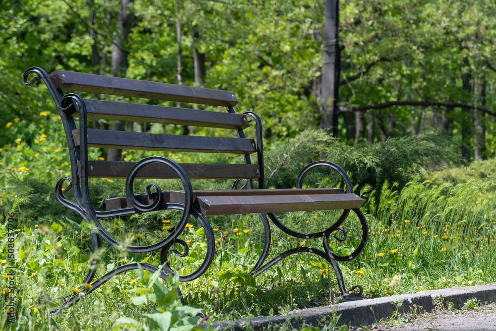 One empty wooden brown bench in the garden. Comfortable seat with metal dark forged details and railings.