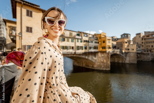 Young woman enjoys beautiful view on famous Old bridge in Florence, sitting on the riverside at sunset. Female traveler visiting italian landmarks. Stylish woman wearing dress and colorful shawl © rh2010