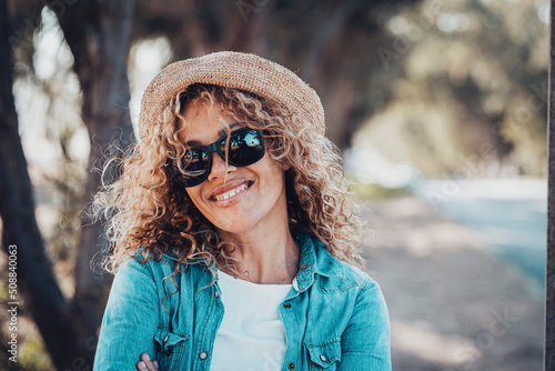 Portrait of young adult woman smiling at the camera with sunglasses and straw hat. Female people happy in outdoors leisure activity. Defocused background with road. Copy space on the right