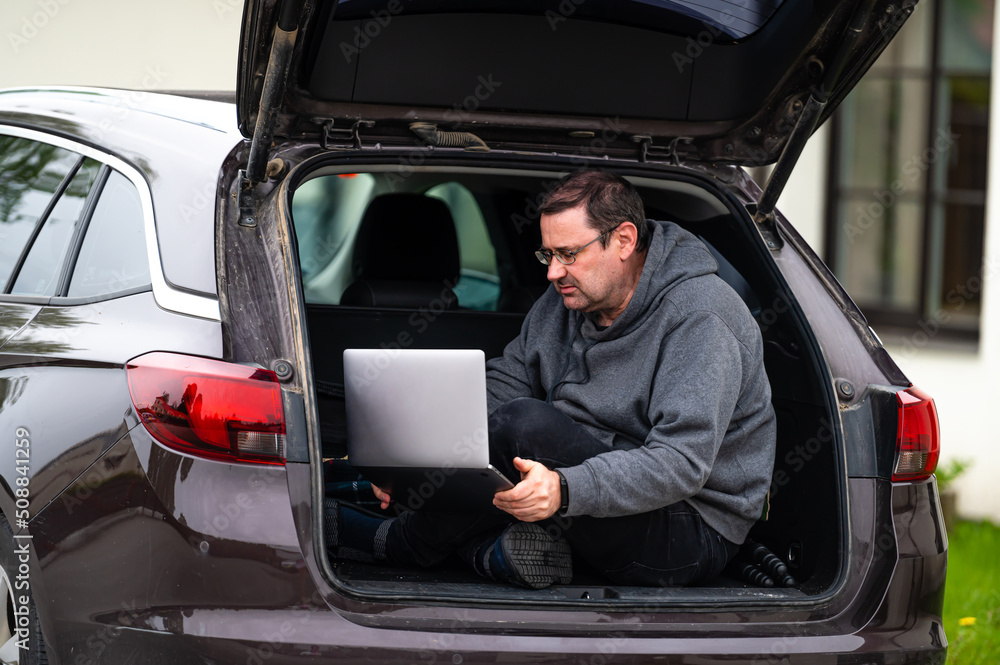 a man sitting in the open trunk of a car and working with a laptop, mobile technology, remote work a man sitting in the open trunk of a car and working with a laptop, mobile technology, remote work