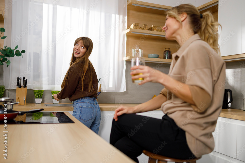 Nice girl washing dishes while her mother is relaxing sitting at the kitchen table with a glass of juice Nice girl washing dishes while her mother is relaxing sitting at the kitchen table with a glass of juice
