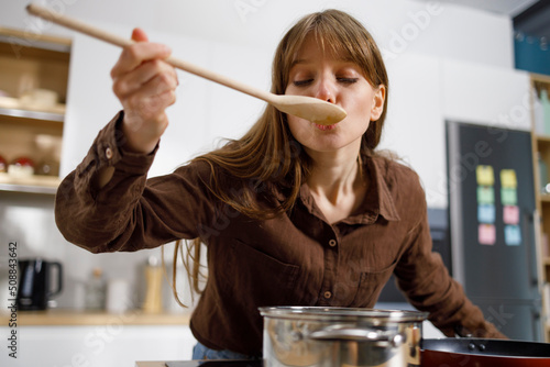 Obraz na plátně Young woman tasting food with wooden spoon while cooking