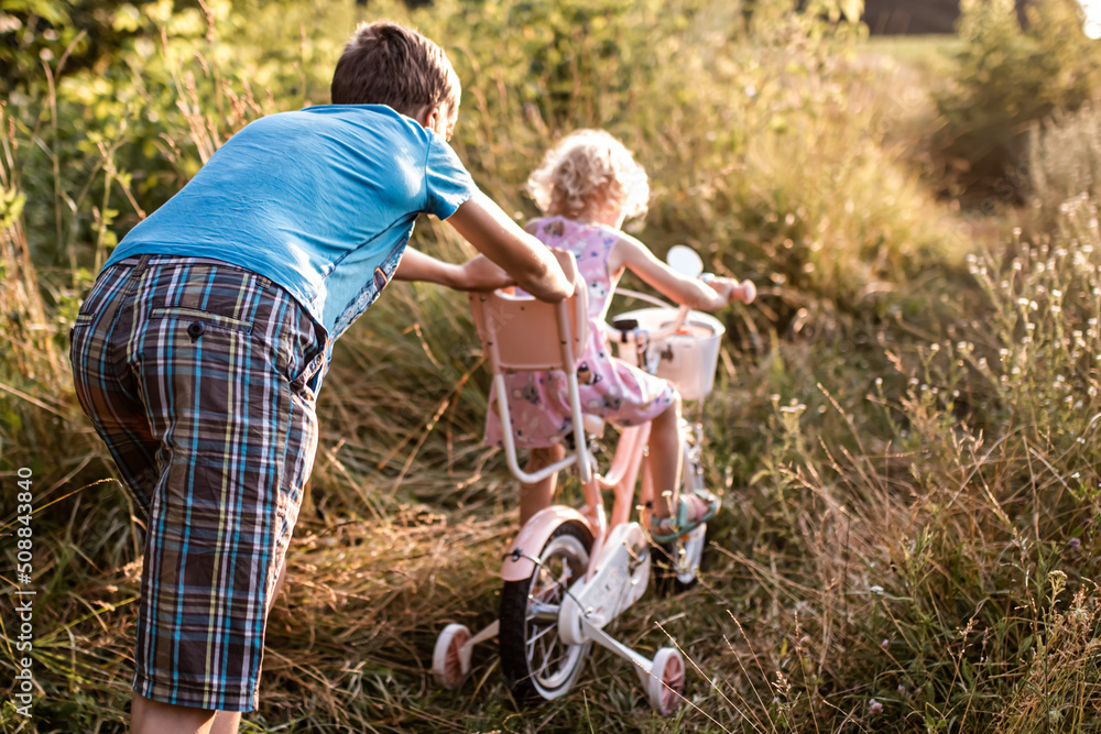brother teaches little sister to ride bicycle. girl rides along rural road on two-wheeled bicycle among summer grasses brother teaches little sister to ride bicycle. girl rides along rural road on two-wheeled bicycle among summer grasses