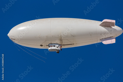 A white blimp without any markings, a blank canvas or banner space with a blue sky in the background. A lighter than air ship flying high with room to put your own ad.