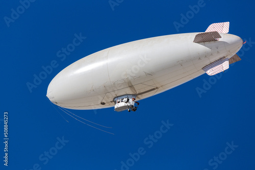 A white blimp without any markings  a blank canvas or banner space with a blue sky in the background. A lighter than air ship flying high with room to put your own ad.