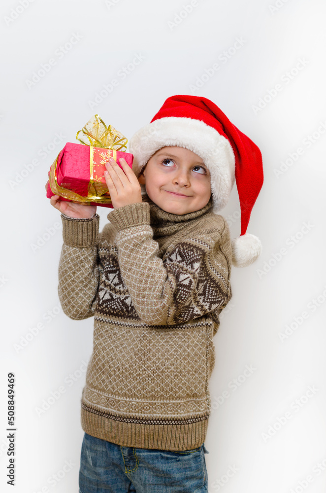 Cute boy in Santa's hat holding a gift and guesses what is in it Cute boy in Santa's hat holding a gift and guesses what is in it