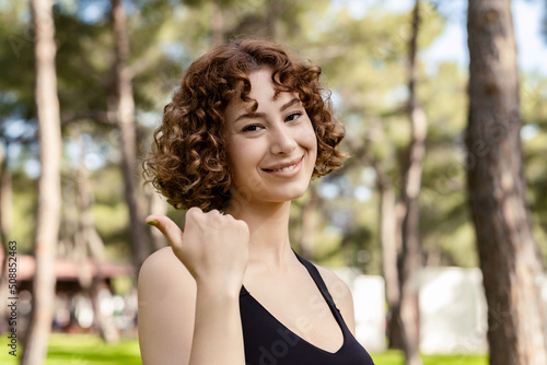Beautiful redhead woman wearing sportive clothes standing on city park, outdoor smiling with happy face looking at the camera and pointing to the side with thumb up. Showing advertisement, indicating.