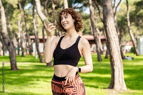 Young redhead girl smiling happy wearing sports bra standing on city park, outdoors using her mobile on speaker phone holding the device in front of face. Happy sportive woman talking with friends.