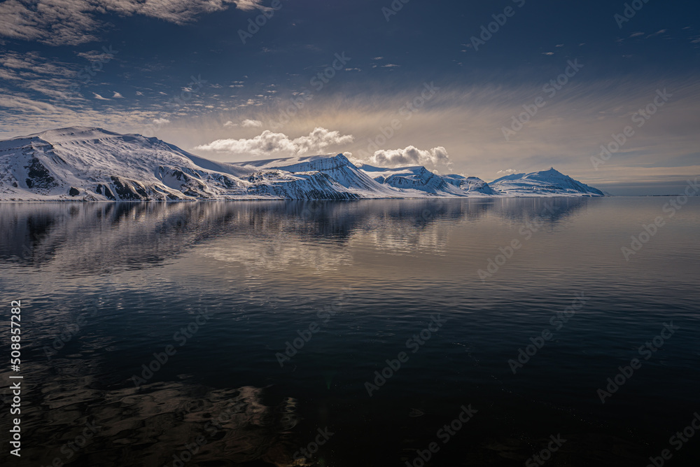 2022-05-15 A ROW OF SNOW COVERED MOUNTAINS WITH A REFLECTION IN THE ARCTIC OCEAN AND NUMEROSU CLOUDS NEAR THE ISLAND OF SVALBARD NORWAY IN THE ARCTIC