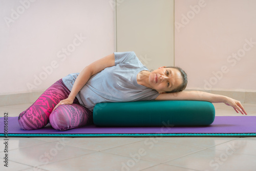 Full length shot of energetic mature woman making exercising and practicing restorative yoga on mat and bolster at home because of social distancing, wearing sports clothes.
