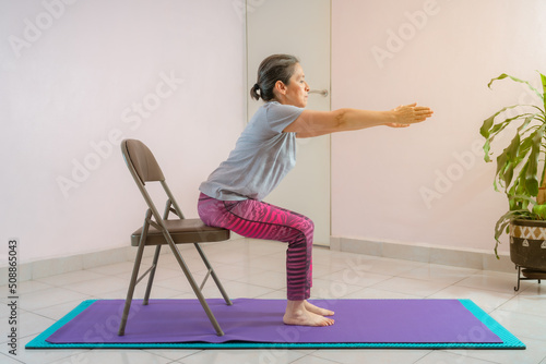 Full length shot of energetic mature woman making exercising and practicing restorative yoga on mat and chair at home because of social distancing, wearing sports clothes.