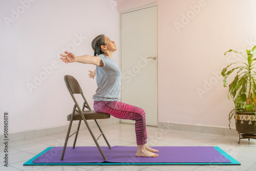 Full length shot of energetic mature woman making exercising and practicing restorative yoga on mat and chair at home because of social distancing, wearing sports clothes.