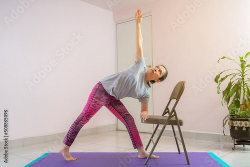 Fotobehang Full length shot of energetic mature woman making exercising and practicing restorative yoga on mat and chair at home because of social distancing, wearing sports clothes