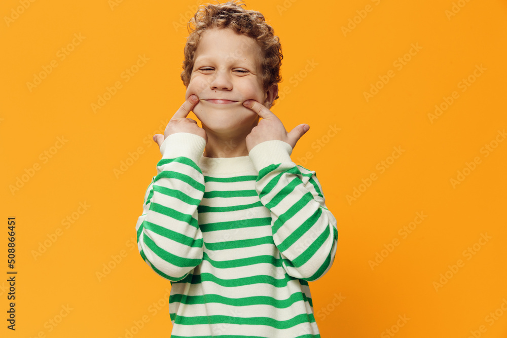 horizontal portrait of a cute, funny, playful, curly-haired boy of school age in a striped long-sleeved t-shirt on an orange background pulling a smile with his fingers