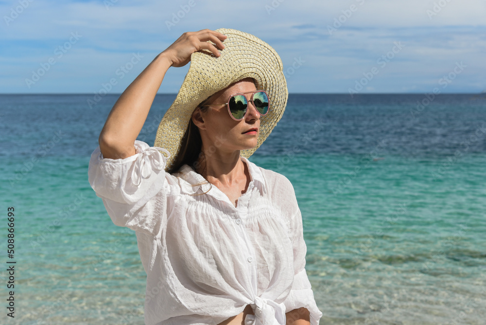 A happy woman is resting on the beach. She is wearing a straw hat, denim shorts, sunglasses and a white shirt. Travel and good rest, happy holidays, tourism, Philippines, summer season, copy space