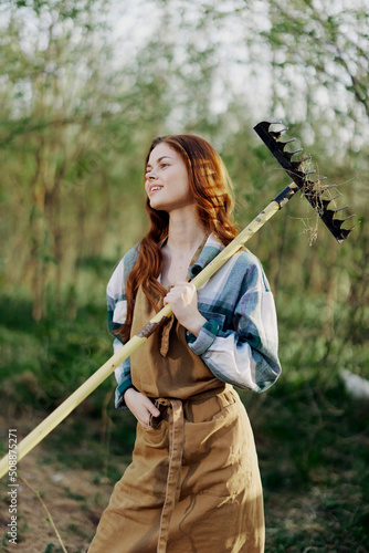 A woman farmer in work clothes and an apron works outdoors in nature and holds a rake to gather grass