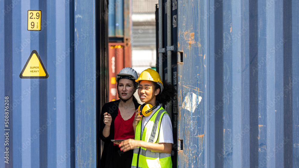 Two female logistic workers from different cultures or ethnicities have conversation next to shipping blue containers. A female foreman discuss with cargo female worker for stocking Two female logistic workers from different cultures or ethnicities have conversation next to shipping blue containers. A female foreman discuss with cargo female worker for stocking