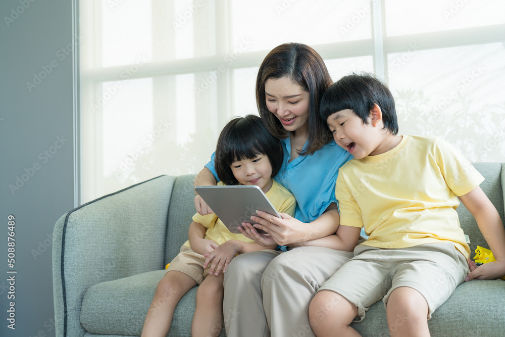 Happy Asian mother teaching two son in tablet while sitting on sofa in living room at home.Family relaxing on holiday. Happy Asian mother teaching two son in tablet while sitting on sofa in living room at home.Family relaxing on holiday.