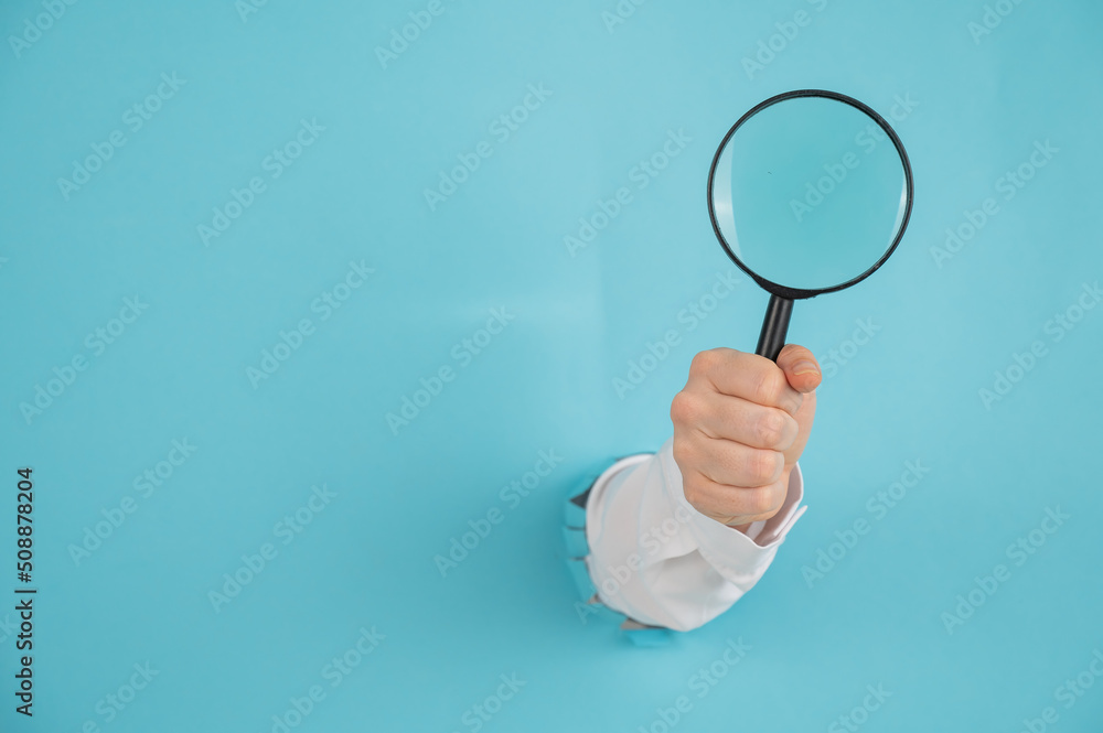 A woman's hand sticking out of a hole in a paper blue background holds a magnifying loupe. A woman's hand sticking out of a hole in a paper blue background holds a magnifying loupe.